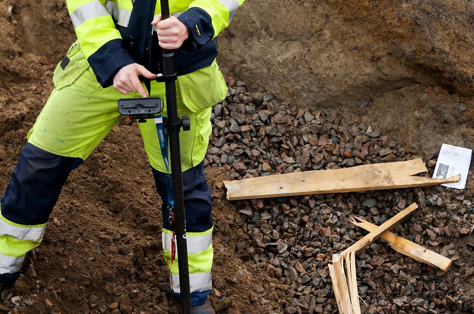 Byggarbetare står i grävschakt med mätinstrument